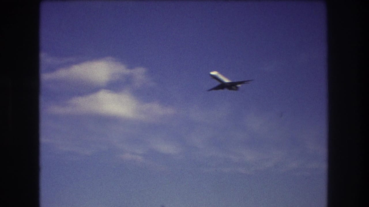 The airplane passes through the right side of the frame near a pale cloud, with saturated 8mm color and visible gate edges from the supplied Boston 1975 clip.