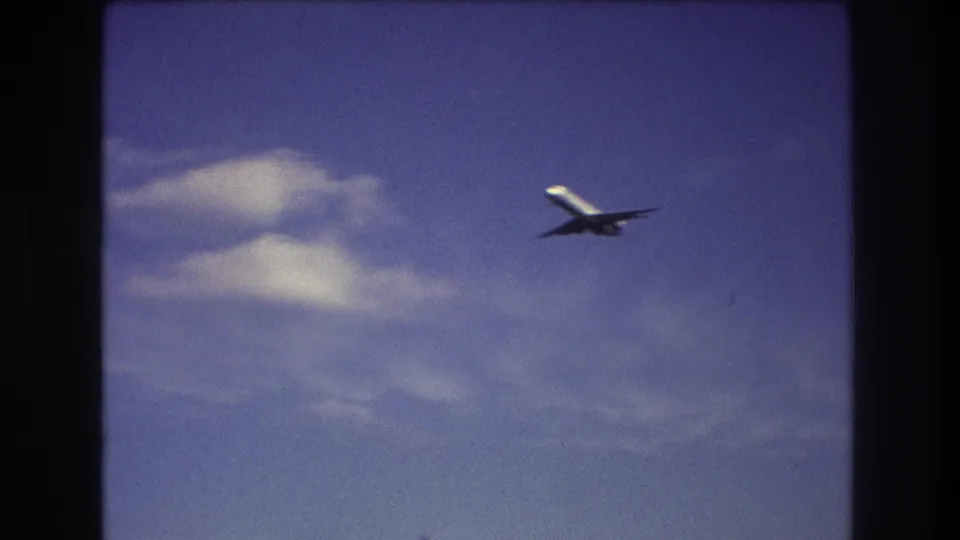 The airplane passes through the right side of the frame near a pale cloud, with saturated 8mm color and visible gate edges from the supplied Boston 1975 clip.