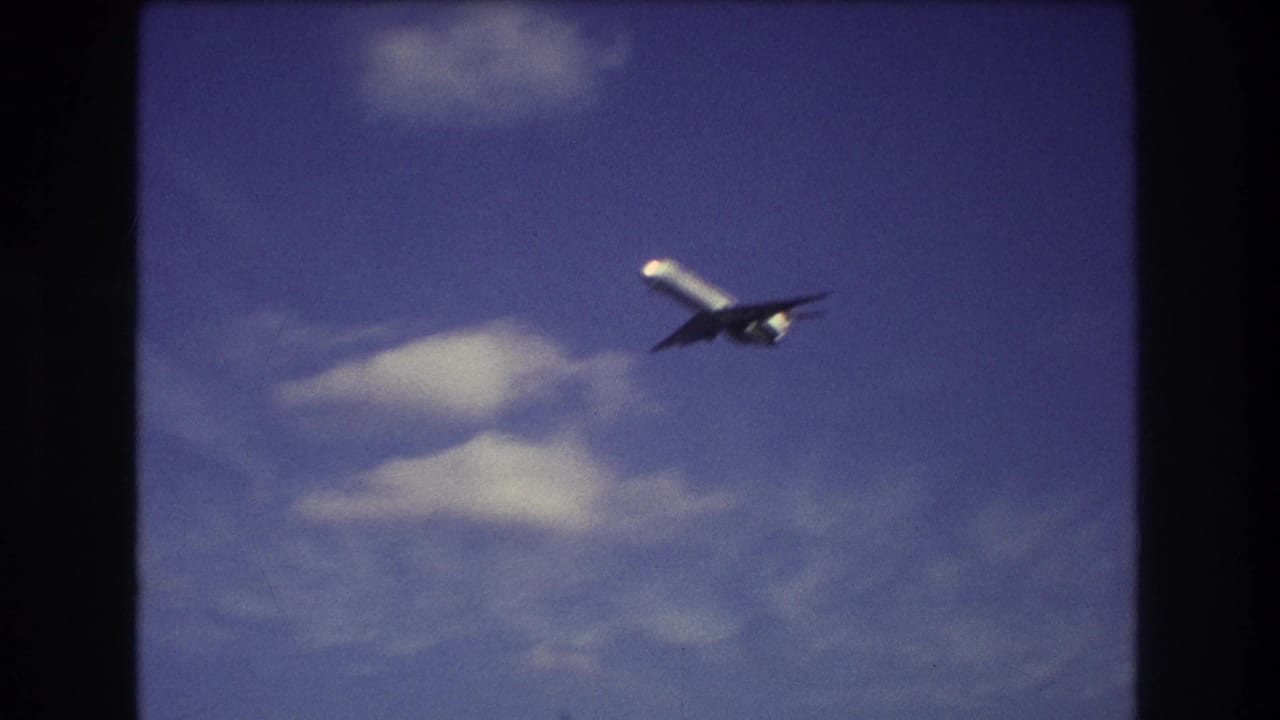 A commercial jet appears high in a blue, partly cloudy sky in 1975 Boston-area 8mm home-movie footage, framed by dark film edges.