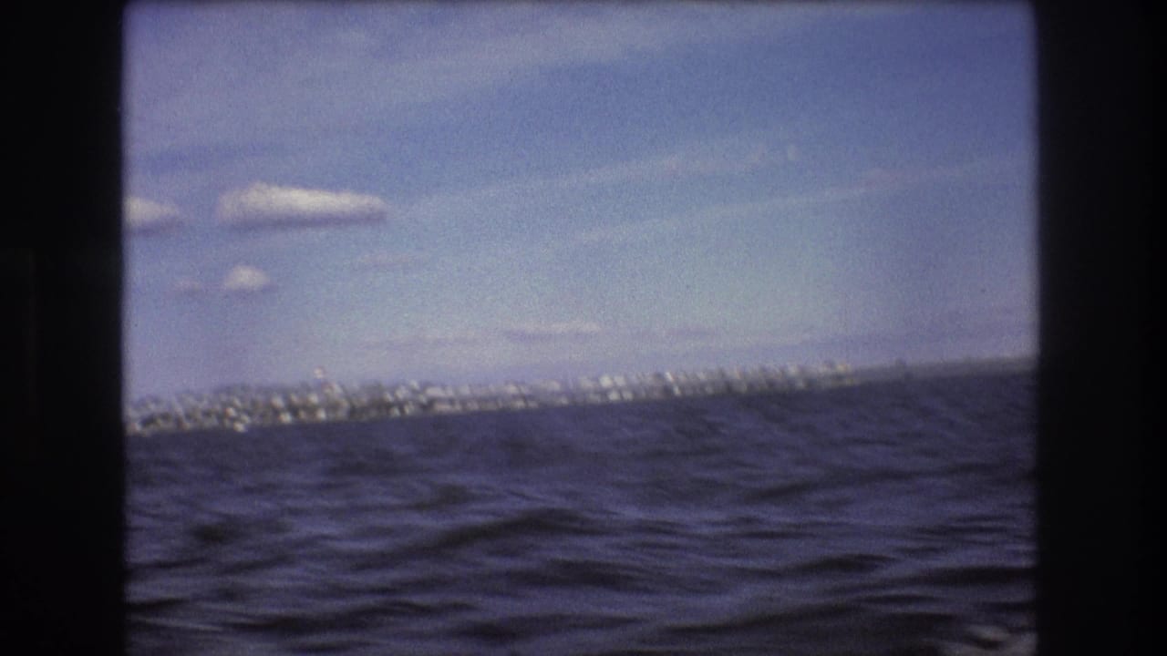 1975 Boston-area 8mm frame from a moving boat showing choppy water, a far shoreline, scattered clouds, and dark gate or window edges.