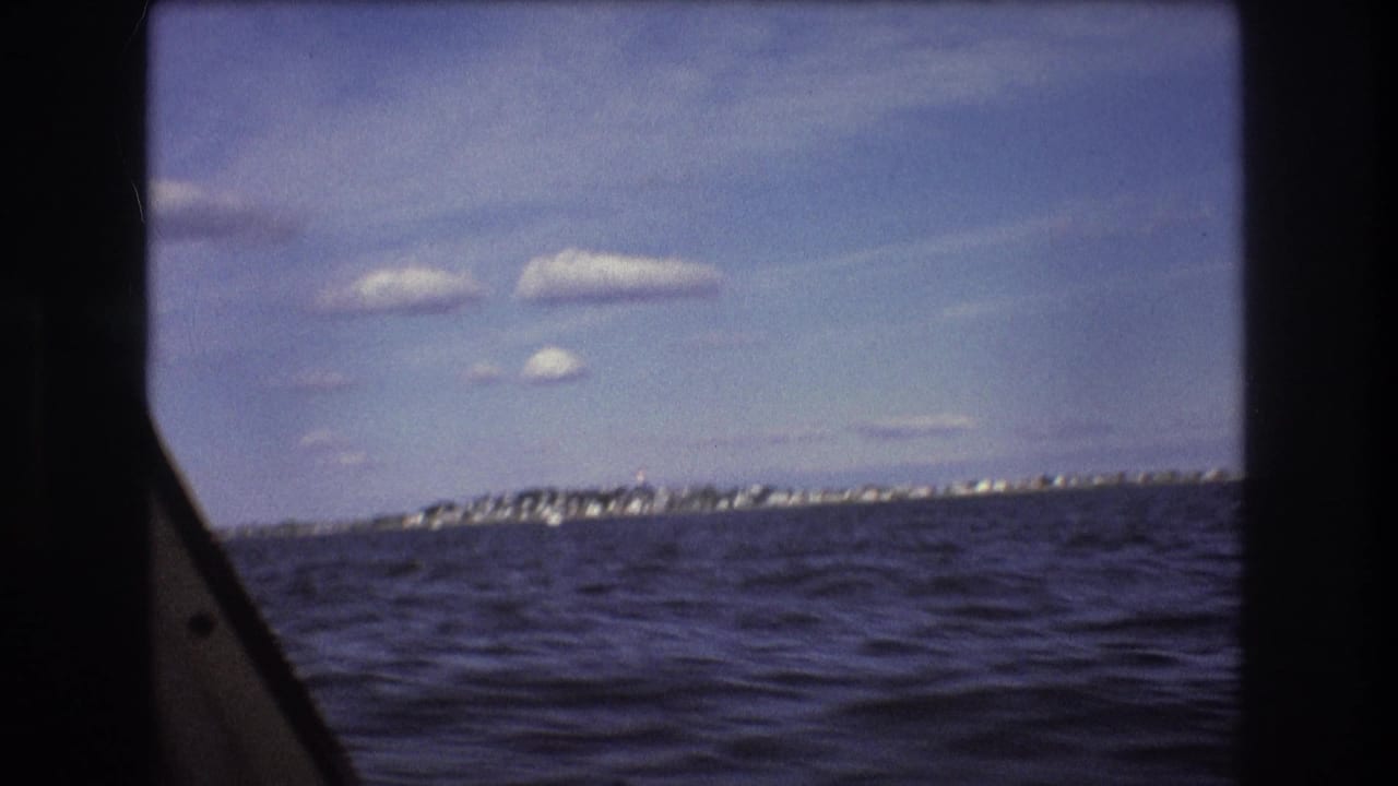 1975 Boston-area boat-view frame with rippling water, distant houses or shoreline structures, scattered clouds, and a dark vertical frame at center.