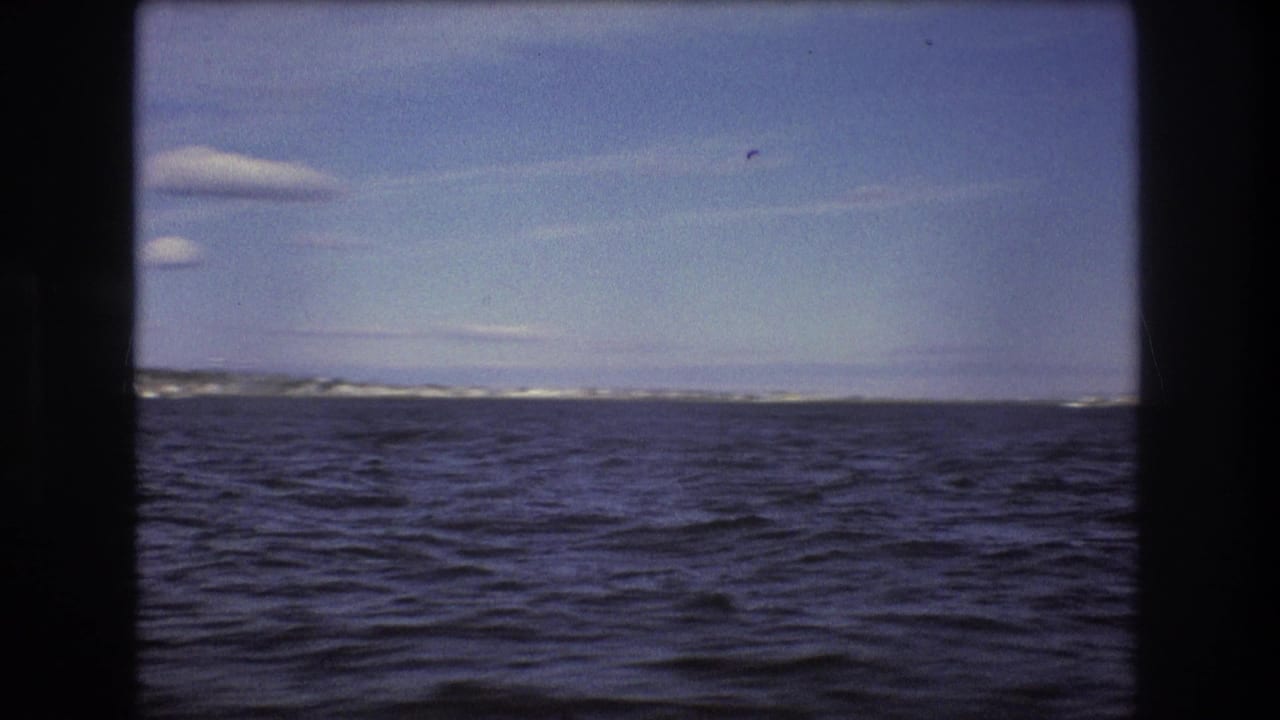 1975 Boston-area 8mm frame showing a wide expanse of choppy water, a low shoreline on the horizon, and a pale sky with small clouds.