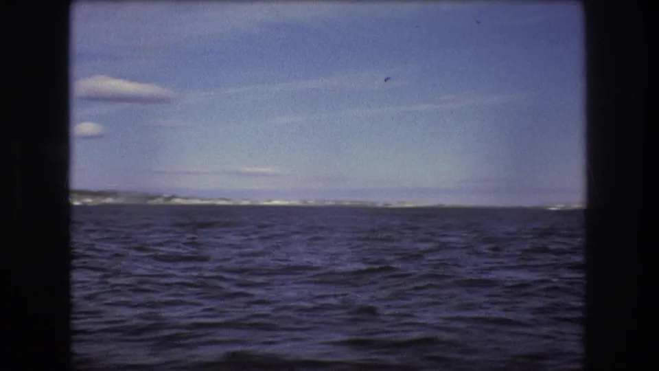 1975 Boston-area 8mm frame showing a wide expanse of choppy water, a low shoreline on the horizon, and a pale sky with small clouds.