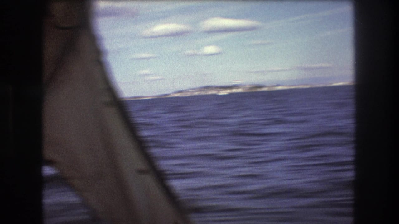 1975 Boston-area home-movie frame looking past a dark boat or window edge across blue water toward a distant shoreline and scattered white clouds.