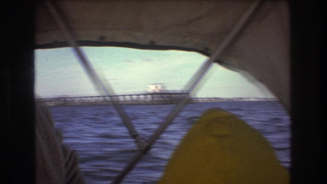 1975 Boston-area 8mm frame from inside a moving boat, showing canopy supports, choppy blue water, and a distant long pier or bridge-like structure.
