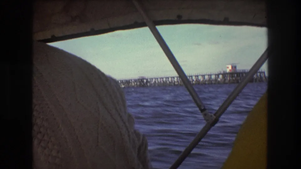 1975 Boston-area boat-view frame with a person in a textured white sweater at left, canopy supports crossing the view, choppy water, and a distant pier-like structure.