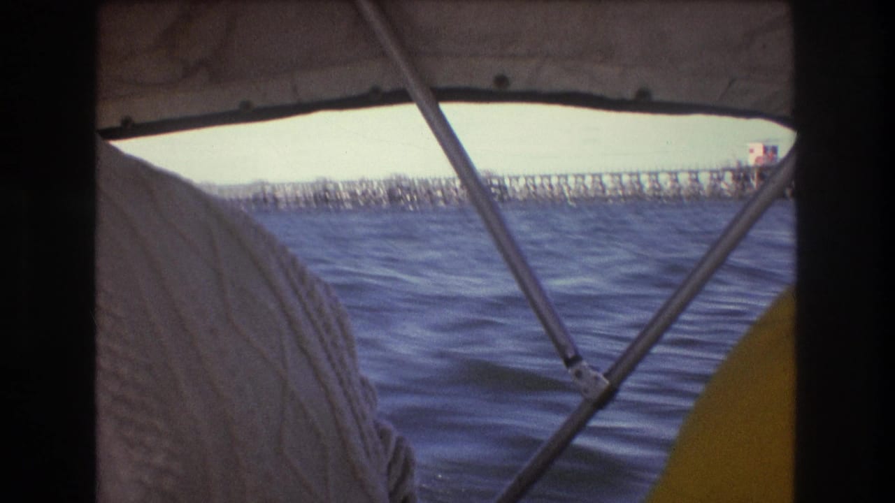 1975 Boston-area 8mm frame from a moving boat, with a sweatered passenger partly blocking the view of choppy water and a long pier-like structure beyond the canopy.