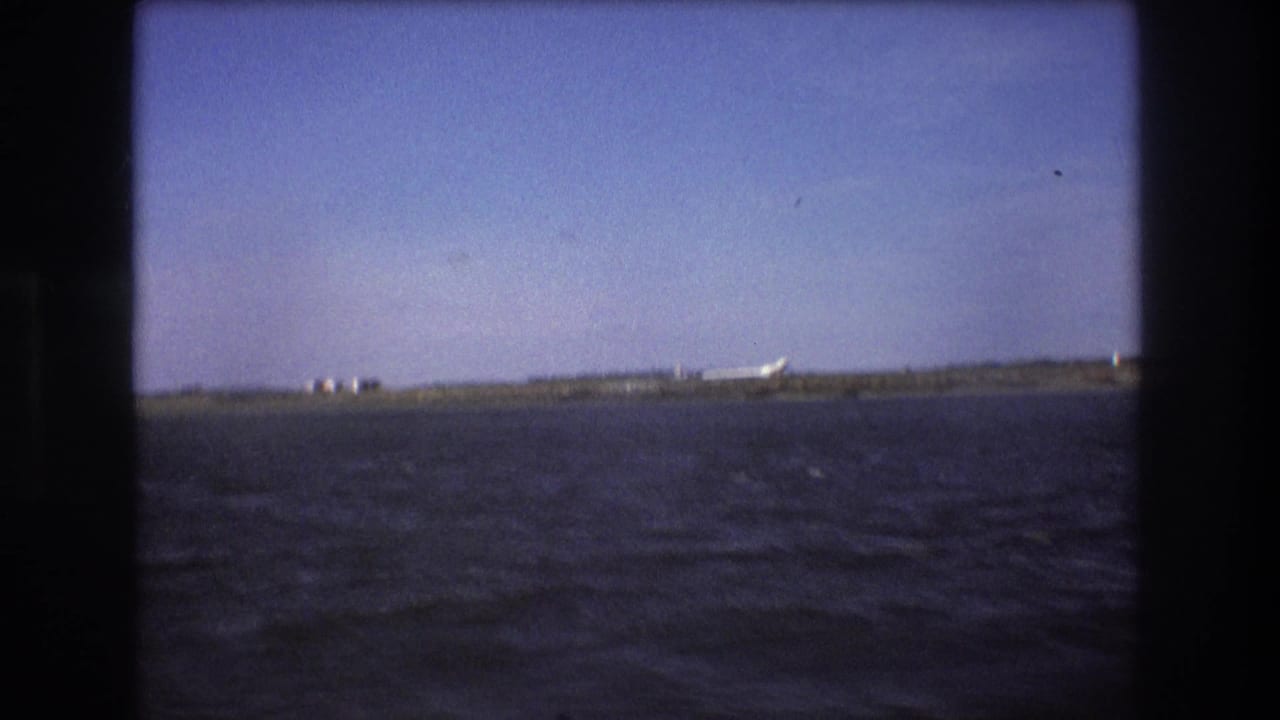 1975 Boston-area 8mm frame looking across dark harbor water toward a distant shoreline where a white airplane appears on the ground.
