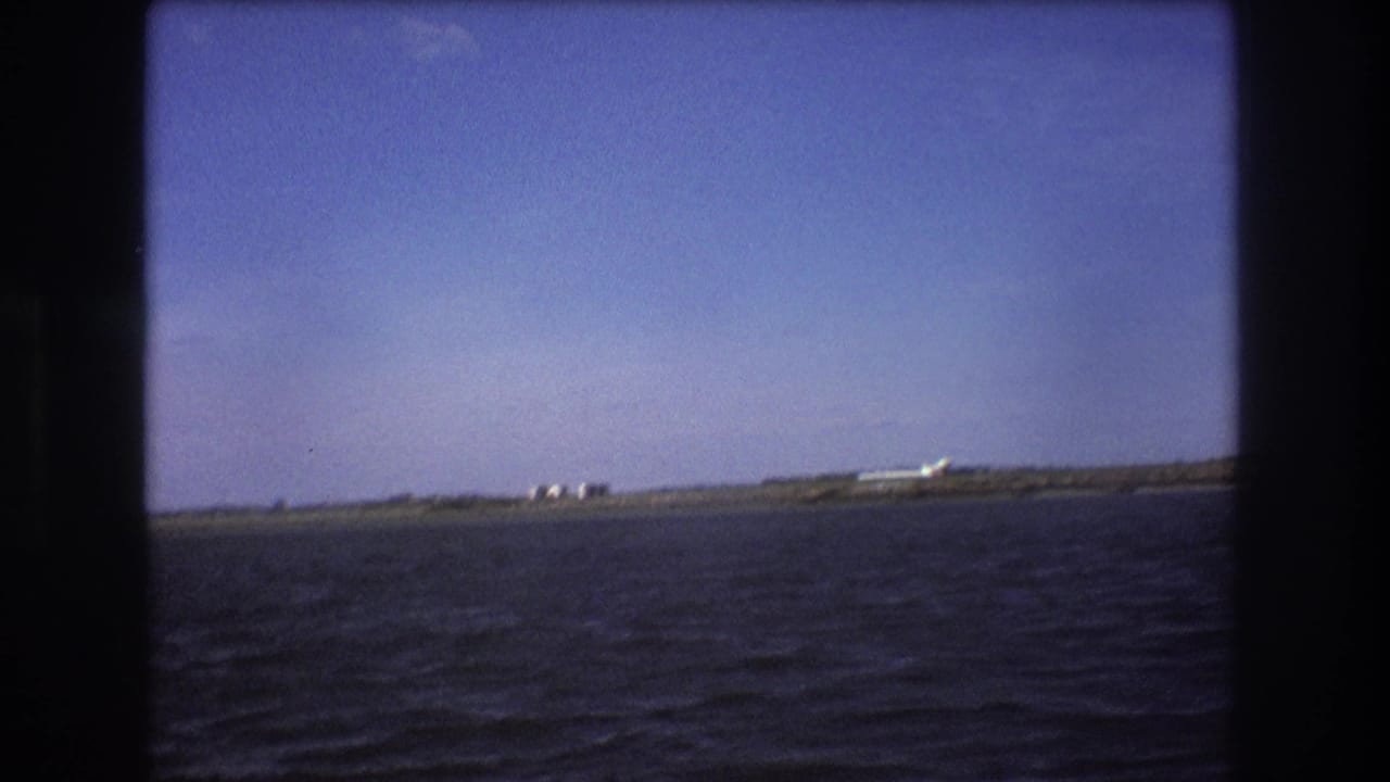 1975 Boston-area 8mm frame showing a distant white airplane near a runway or shoreline beyond dark rippled water under blue sky.