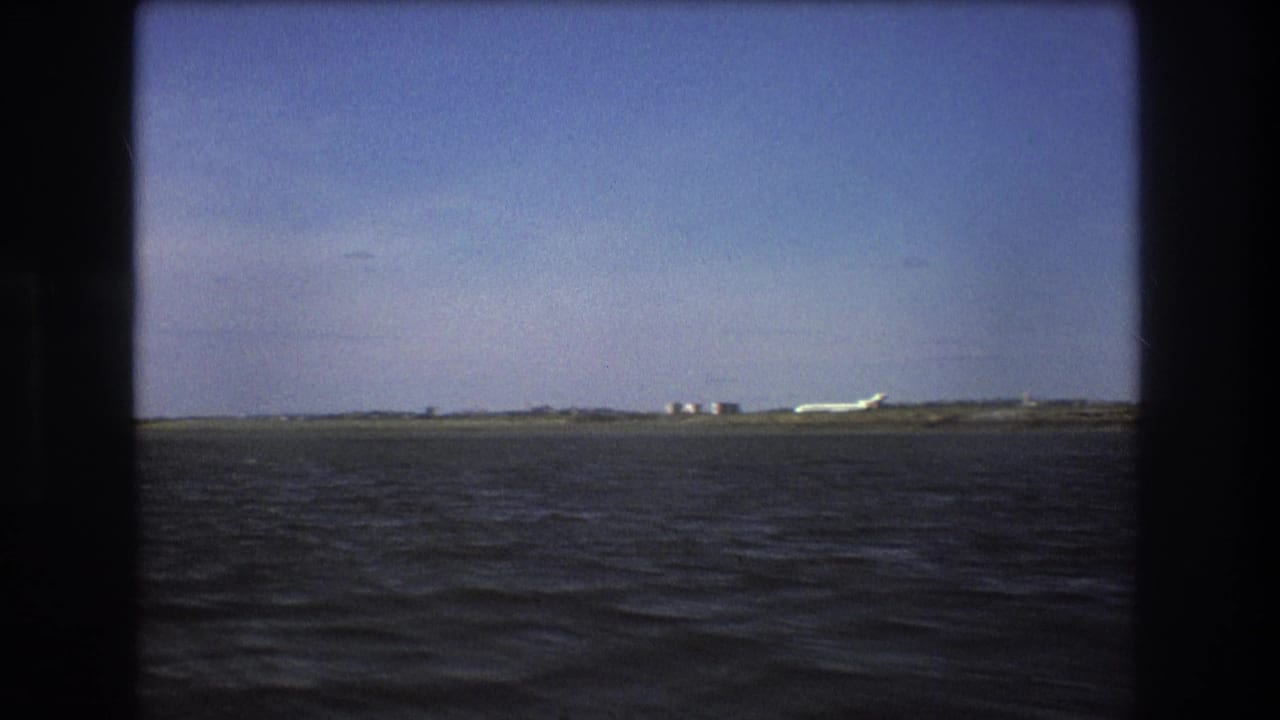 1975 Boston-area 8mm view of a white aircraft on a distant runway-like strip, seen across open water with a clear sky above.