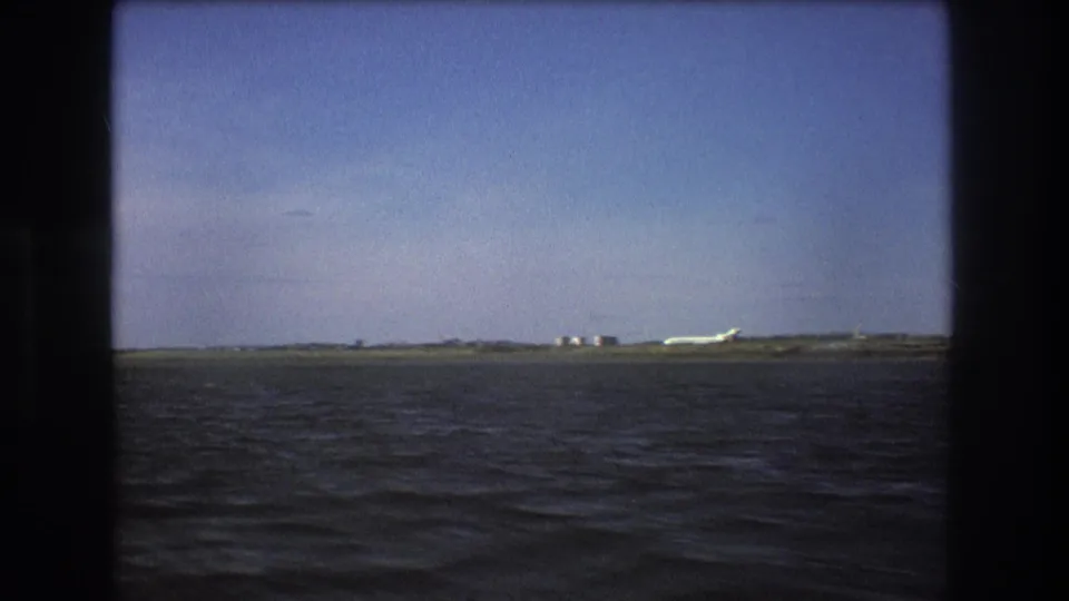 1975 Boston-area 8mm view of a white aircraft on a distant runway-like strip, seen across open water with a clear sky above.