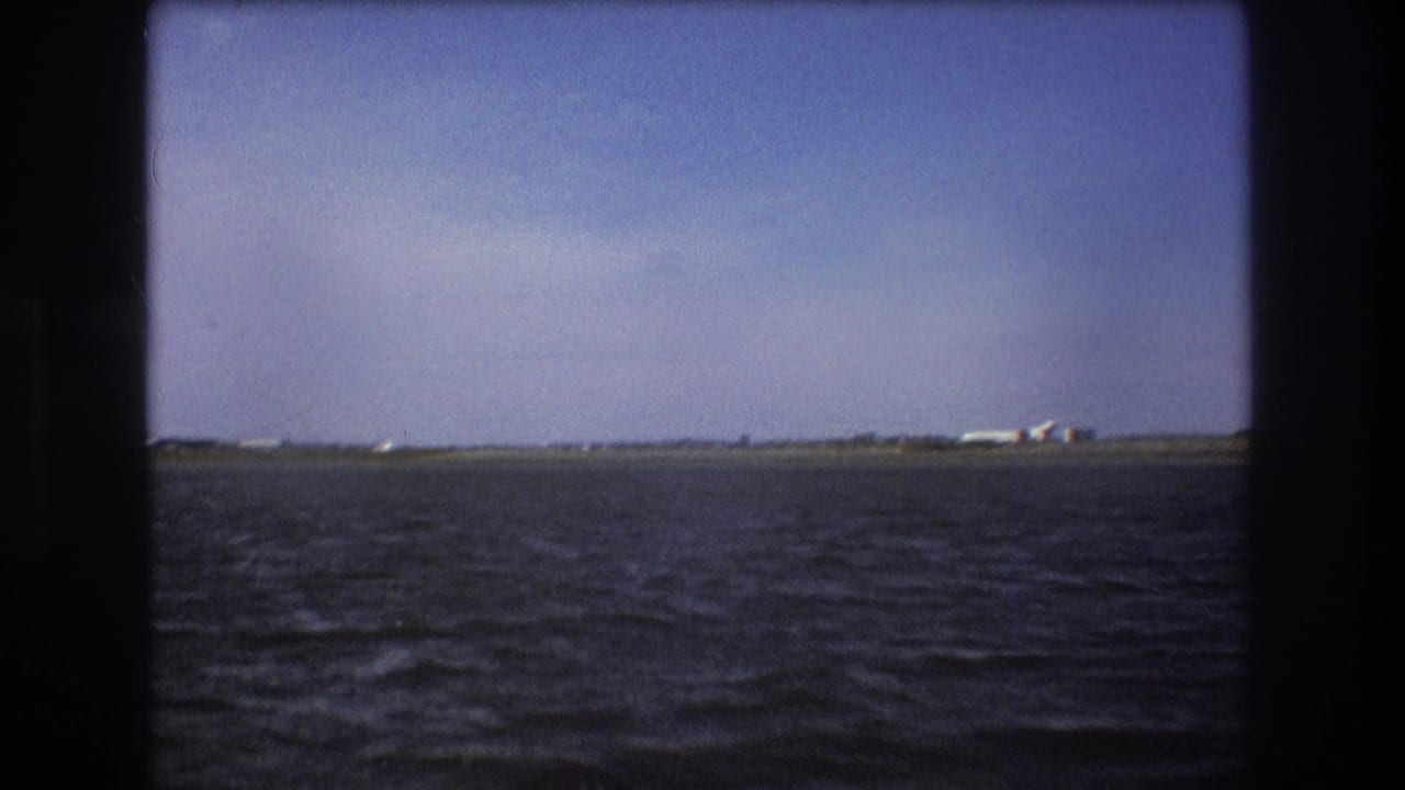 1975 Boston-area home-movie frame with choppy water in the foreground, a low runway or shoreline, and a distant white airplane near small structures.