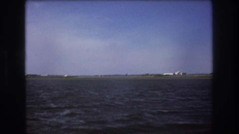 1975 Boston-area home-movie frame with choppy water in the foreground, a low runway or shoreline, and a distant white airplane near small structures.