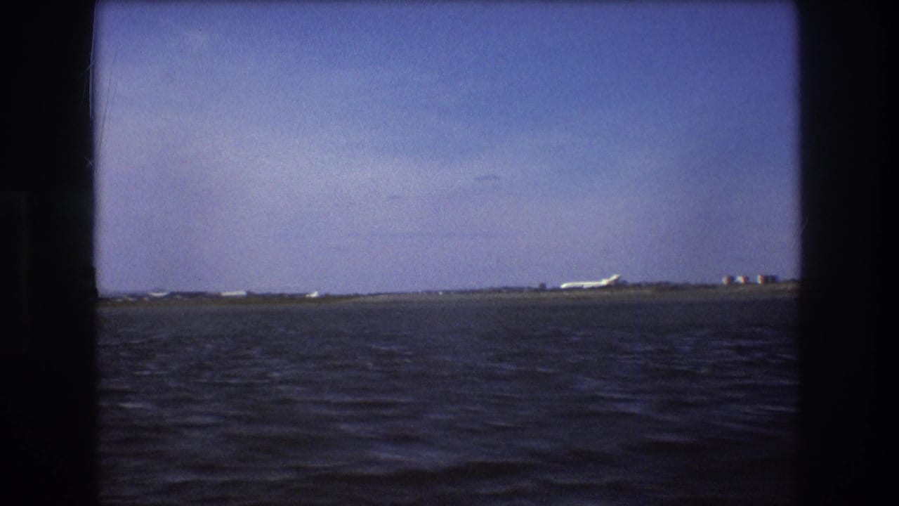 1975 Boston-area 8mm frame showing a distant white aircraft near the right side of a low coastal runway, with rippled water and blue sky dominating the view.