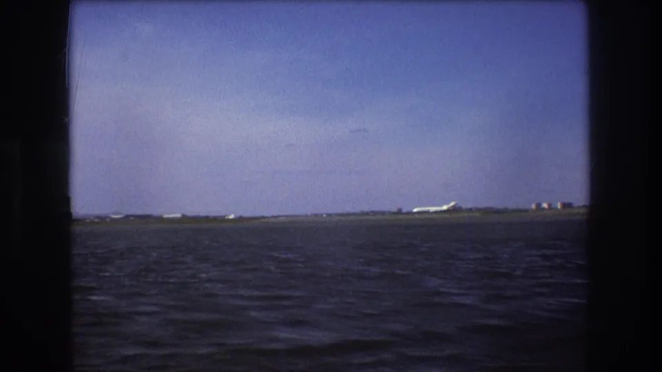 1975 Boston-area 8mm frame showing a distant white aircraft near the right side of a low coastal runway, with rippled water and blue sky dominating the view.
