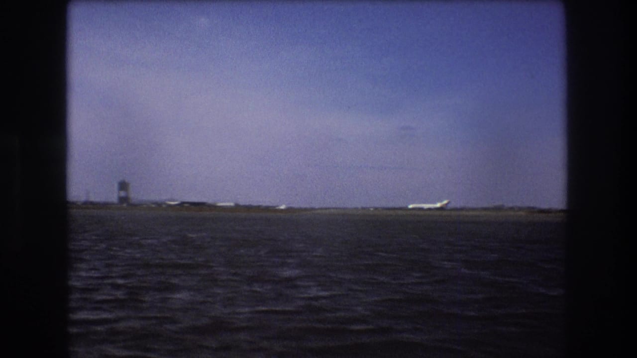 Boston 1975 home-movie frame showing choppy water in the foreground, a distant airport shoreline, a tower, and a white aircraft near the runway under hazy sky.