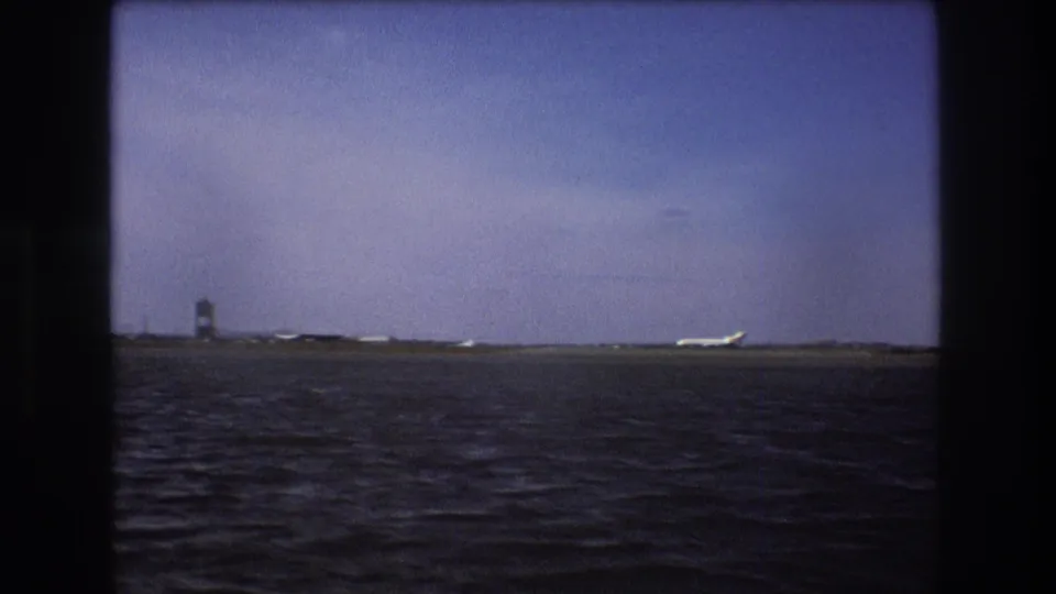 Boston 1975 home-movie frame showing choppy water in the foreground, a distant airport shoreline, a tower, and a white aircraft near the runway under hazy sky.
