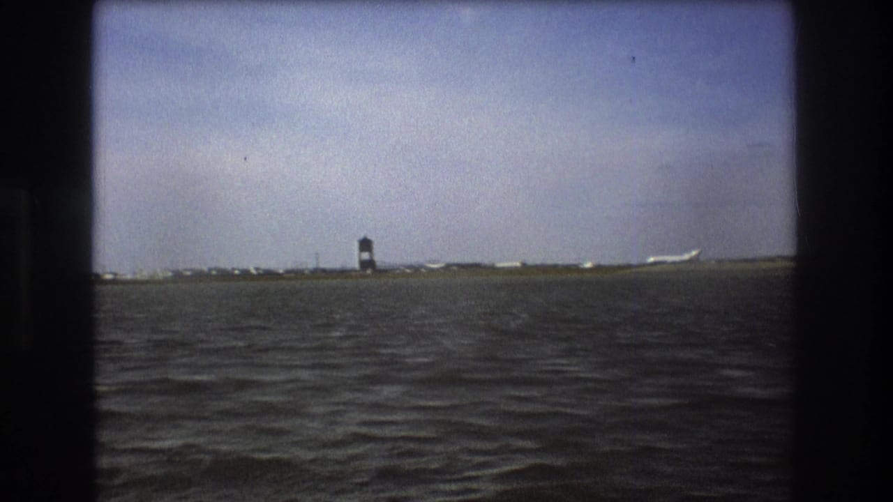Boston 1975 frame with rippling water, a low airport horizon, a dark control tower, and a white passenger aircraft seen at a distance.