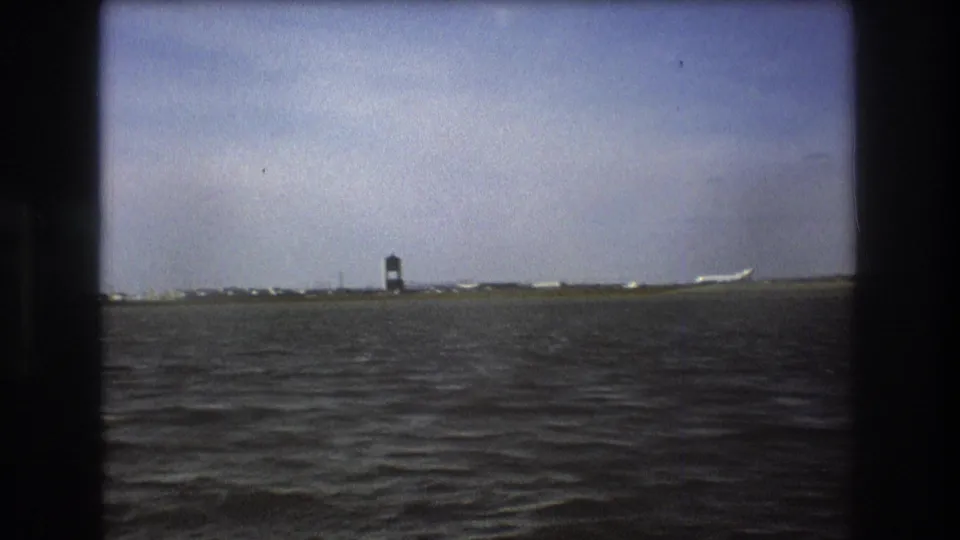 Boston 1975 frame with rippling water, a low airport horizon, a dark control tower, and a white passenger aircraft seen at a distance.