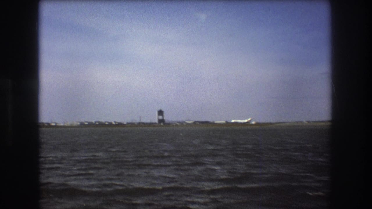 Boston 1975 8mm still showing dark choppy water, a distant control tower, runway buildings, and a white aircraft near the horizon.