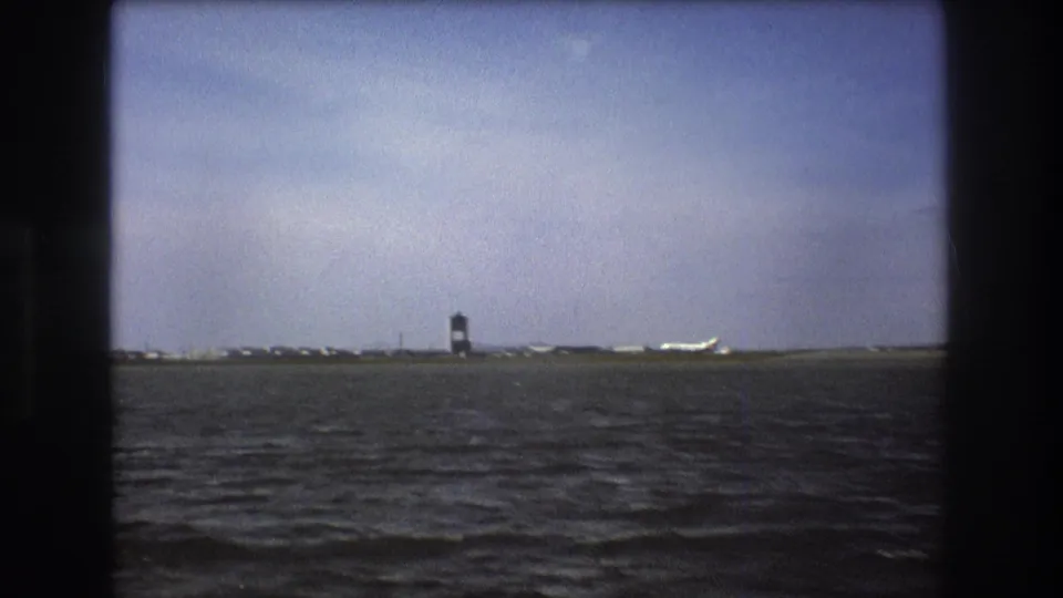 Boston 1975 8mm still showing dark choppy water, a distant control tower, runway buildings, and a white aircraft near the horizon.