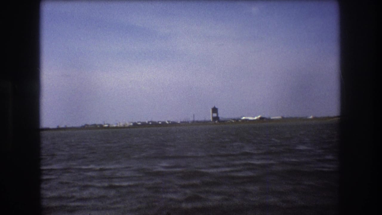 Boston 1975 home-movie frame with the airport tower, a white aircraft near the runway, low shoreline structures, and choppy foreground water framed by dark 8mm edges.