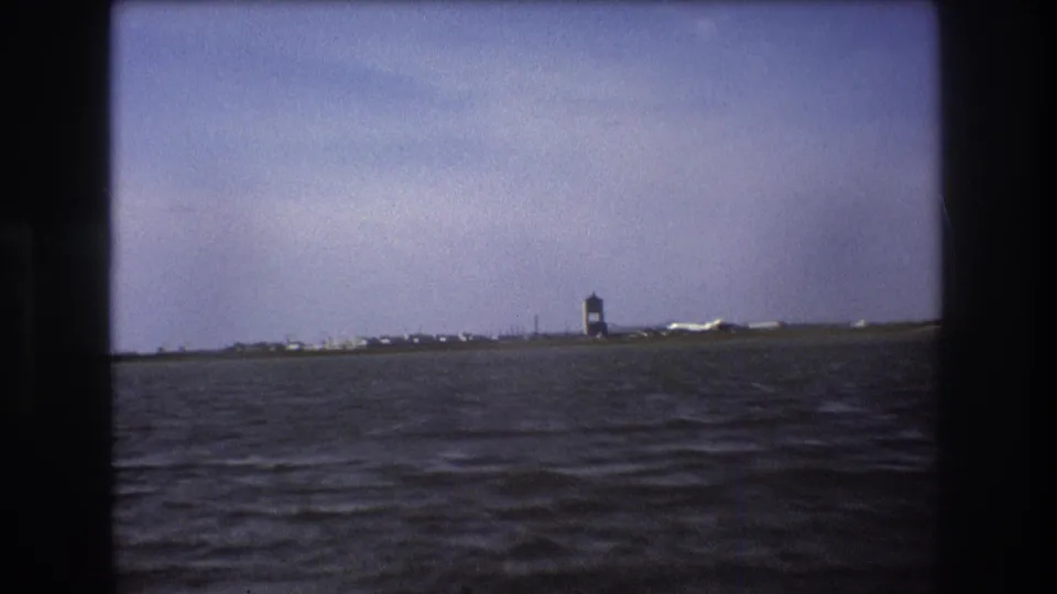 Boston 1975 home-movie frame with the airport tower, a white aircraft near the runway, low shoreline structures, and choppy foreground water framed by dark 8mm edges.