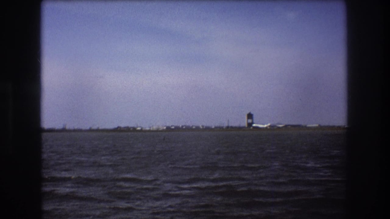 1975 Boston-area 8mm frame showing rippled water in the foreground and a distant shoreline with a tower-like structure and low airport-side buildings.