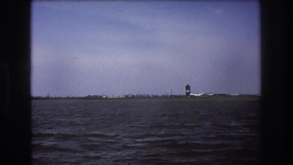1975 Boston-area 8mm frame showing rippled water in the foreground and a distant shoreline with a tower-like structure and low airport-side buildings.
