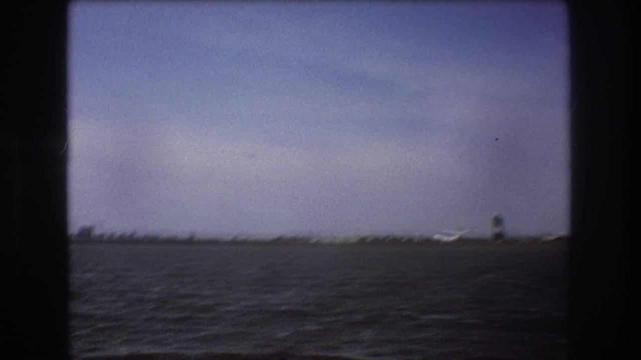 1975 Boston-area 8mm frame looking across water toward a coastal runway, with a small aircraft visible near the shoreline.