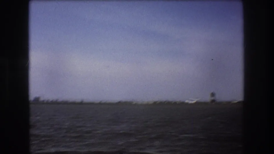 1975 Boston-area 8mm frame looking across water toward a coastal runway, with a small aircraft visible near the shoreline.