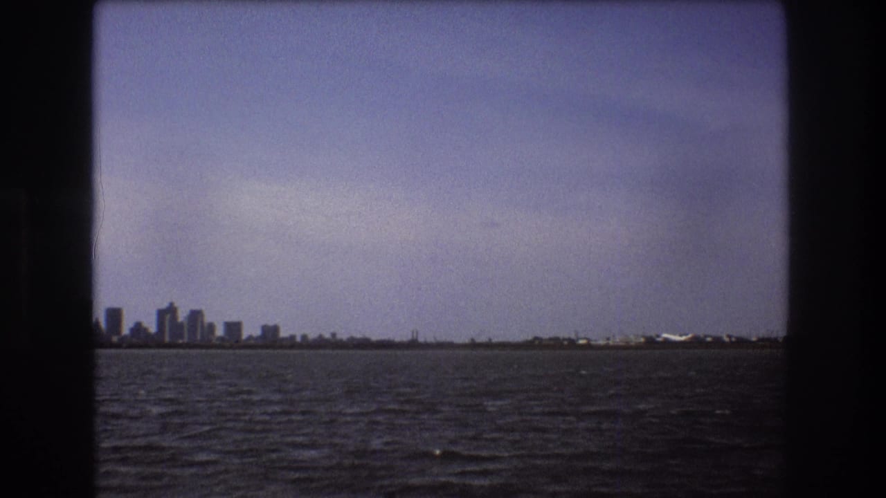 1975 Boston-area 8mm frame showing a distant city skyline and shoreline across dark rippled water from a moving boat viewpoint.