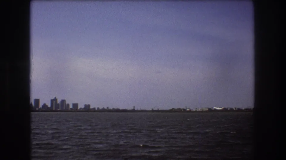 1975 Boston-area 8mm frame showing a distant city skyline and shoreline across dark rippled water from a moving boat viewpoint.