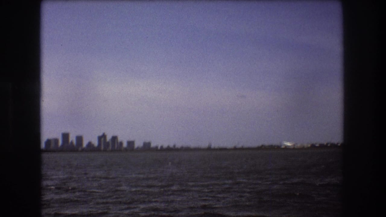 1975 Boston-area 8mm frame with the city skyline to the left and a small aircraft near the distant coastal runway across open water.