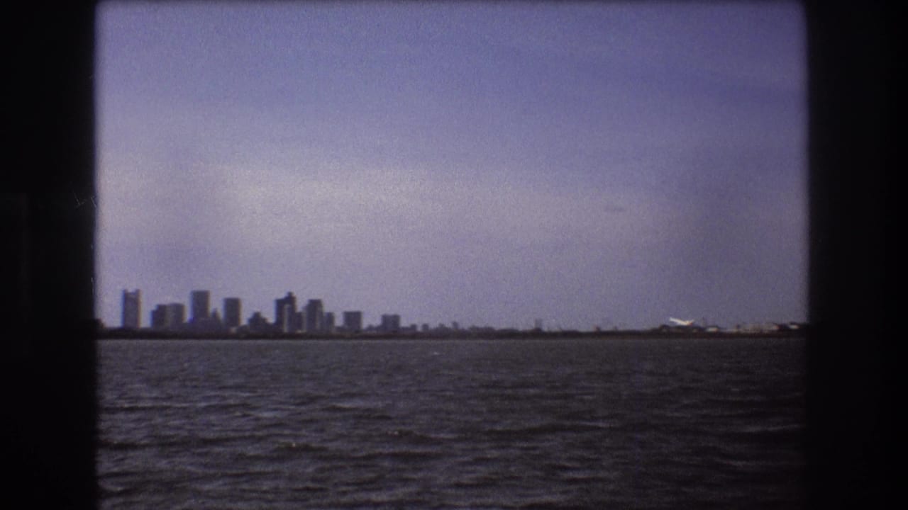 1975 Boston home-movie frame showing a distant skyline across dark rippled water under a hazy blue-purple sky, with the 8mm film gate visible around the view.