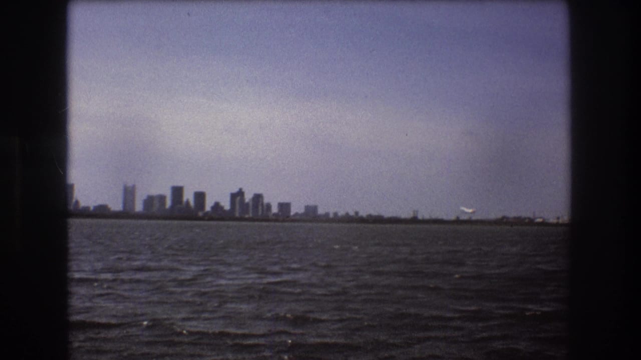 1975 Boston waterfront view with the skyline in the distance and a small white aircraft lifting near the right side of the horizon, framed by dark 8mm borders.