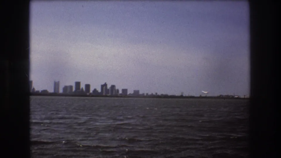 1975 Boston waterfront view with the skyline in the distance and a small white aircraft lifting near the right side of the horizon, framed by dark 8mm borders.