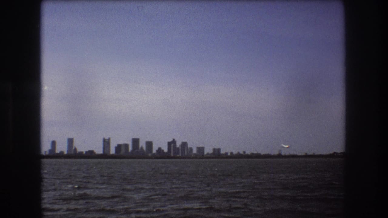 Soft 8mm frame of the Boston skyline across the water in 1975, with a small aircraft visible near the right horizon against the pale sky.