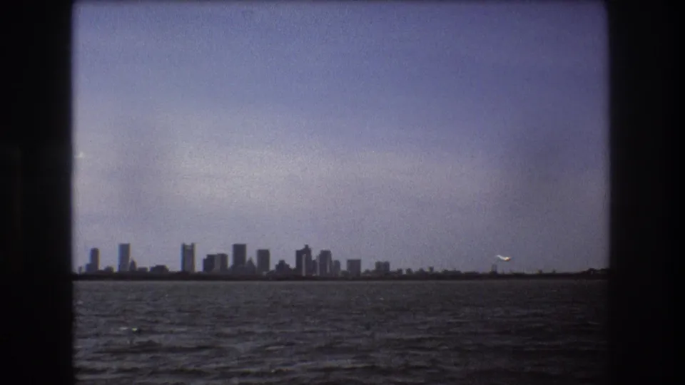 Soft 8mm frame of the Boston skyline across the water in 1975, with a small aircraft visible near the right horizon against the pale sky.
