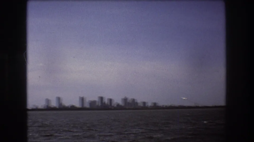 A later 1975 Boston home-movie frame shows the skyline softened by distance and film focus, with rippled water below and a small airplane near the right horizon.
