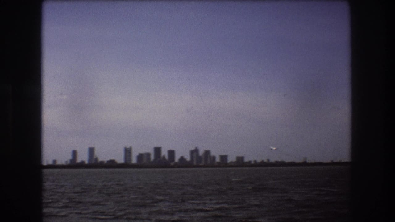 Distant Boston skyline across water in 1975 home-movie footage, framed by dark vertical and horizontal borders with a small airplane in the sky.
