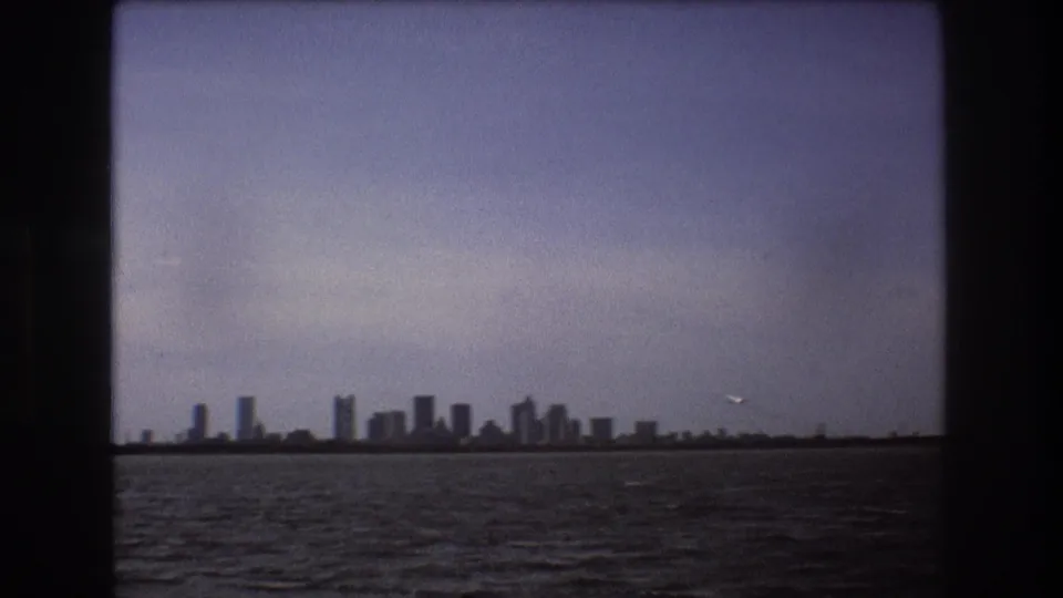 Distant Boston skyline across water in 1975 home-movie footage, framed by dark vertical and horizontal borders with a small airplane in the sky.
