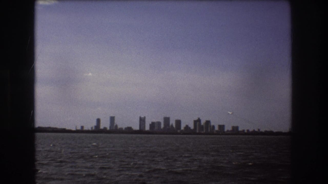 Boston skyline appears low on the horizon across rippled water in 1975 8mm footage, with muted color and heavy dark borders.