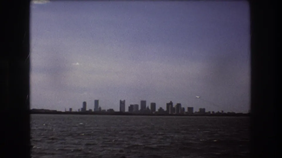 Boston skyline appears low on the horizon across rippled water in 1975 8mm footage, with muted color and heavy dark borders.