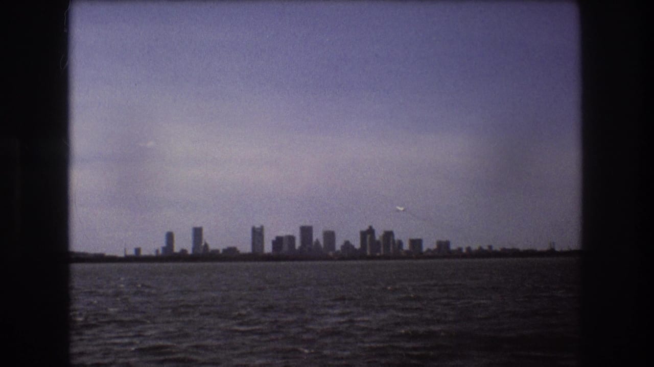Distant 1975 Boston skyline across water with a small white airplane in the cloudy sky and dark borders from the filmed viewpoint.