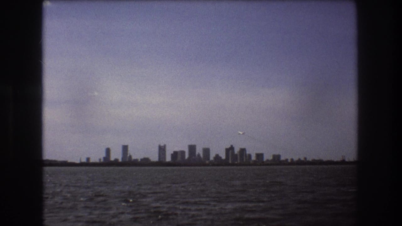 Distant Boston skyline seen across harbor water in 1975 8mm footage, with a small airplane passing above the waterfront.