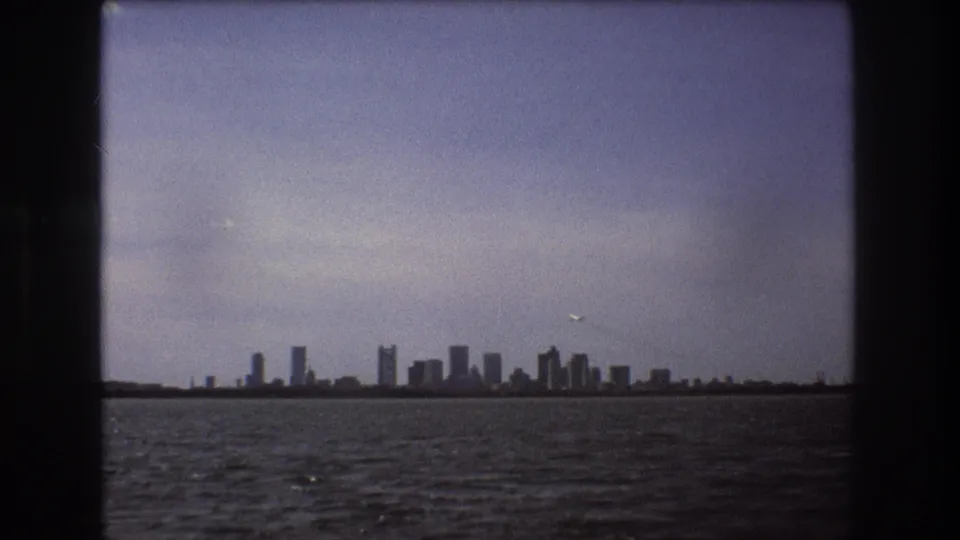 Distant Boston skyline seen across harbor water in 1975 8mm footage, with a small airplane passing above the waterfront.