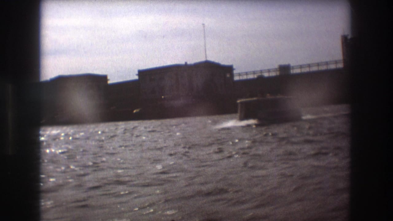 1975 Boston home-movie frame showing choppy harbor water, a low boat, and waterfront buildings under a pale overcast sky.