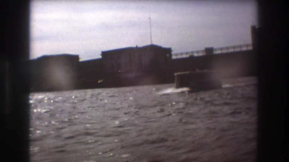 1975 Boston home-movie frame showing choppy harbor water, a low boat, and waterfront buildings under a pale overcast sky.