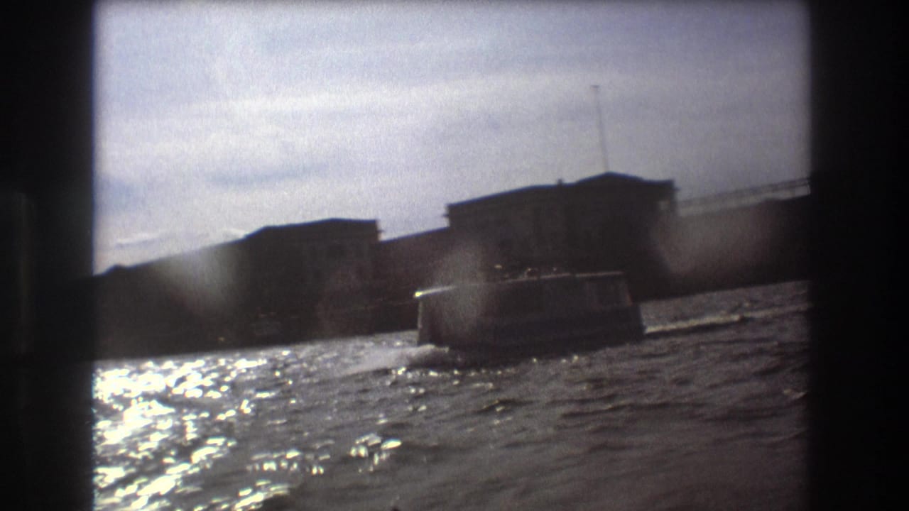 1975 Boston waterfront view from a moving boat, with another small craft crossing rippled water in front of dark shoreline buildings.